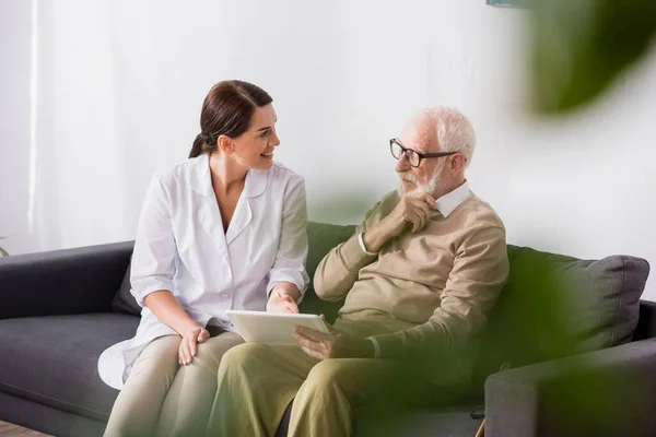 Lächelnder Sozialarbeiter zeigt auf digitales Tablet neben nachdenklichem alten Mann im verschwommenen Vordergrund — Stockfoto