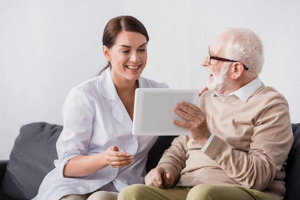 Happy aged man showing digital tablet to excited social worker — Stock Photo