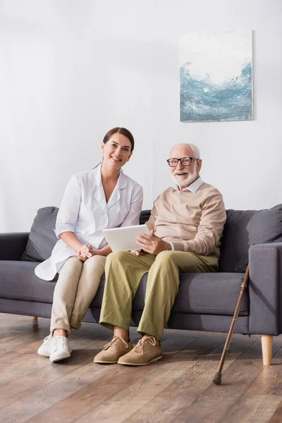 Alegre anciano hombre sosteniendo tableta digital cerca sonriente trabajador social mientras se sienta o sofá en casa - foto de stock