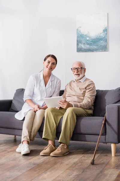 Homme âgé gai tenant tablette numérique près de travailleur social souriant tout en étant assis o canapé à la maison — Photo de stock