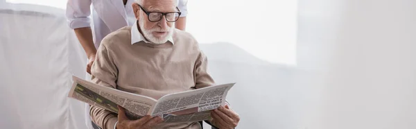 Aged man in eyeglasses reading newspaper near social worker at home, banner — Stock Photo