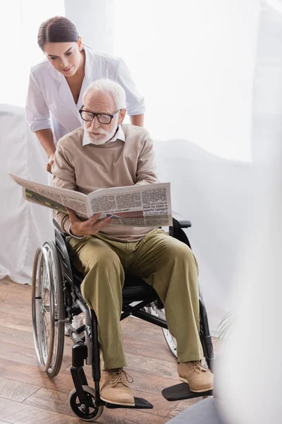 Aged disabled man reading newspaper in wheelchair near social worker — Stock Photo