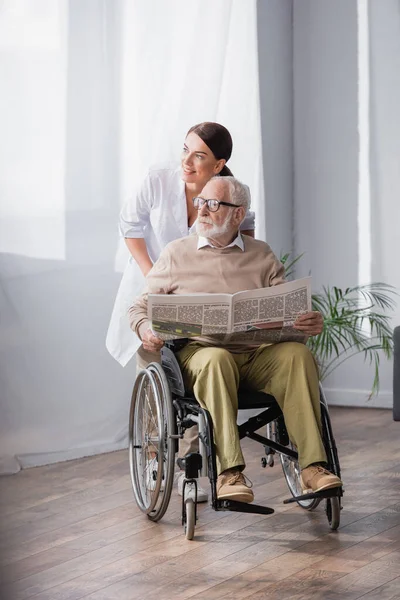 Enfermera geriátrica y hombre discapacitado con periódico mirando a casa - foto de stock