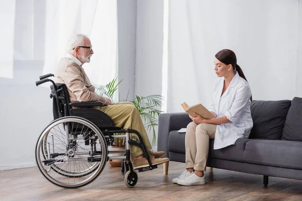 Trabajador social leyendo libro a hombre discapacitado en silla de ruedas en casa — Stock Photo