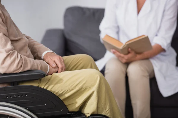 Cropped view of geriatric nurse reading book to elderly man at home — Stock Photo