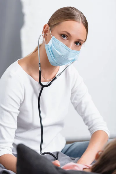 Selective focus of woman in medical mask using stethoscope near child at home — Stock Photo