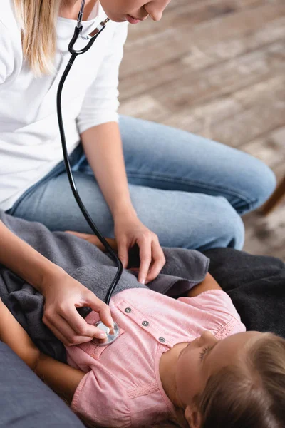 Selective focus of young woman holding stethoscope near chest of daughter during illness at home — Stock Photo