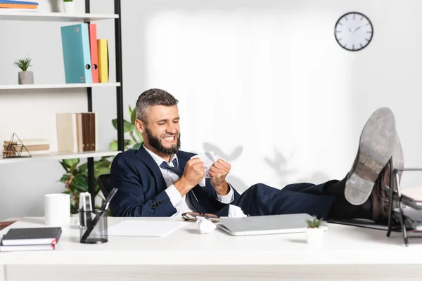 Empresario estresado sosteniendo papel agrupado cerca de la computadora portátil y artículos de papelería en primer plano borroso - foto de stock