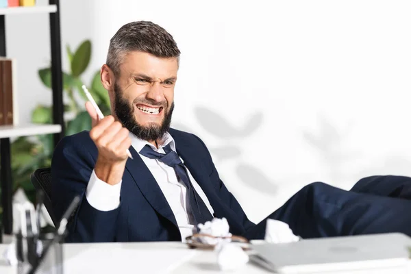 Hombre de negocios nervioso sosteniendo lápiz cerca de papel agrupado y portátil en primer plano borroso en la oficina - foto de stock