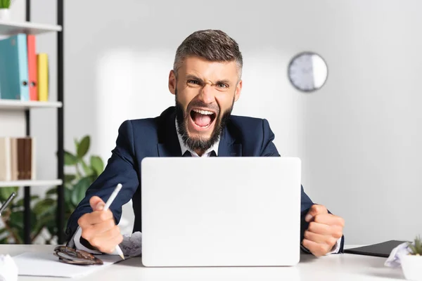 Angry businessman screaming while holding broken pencil near laptop on blurred foreground in office — Stock Photo