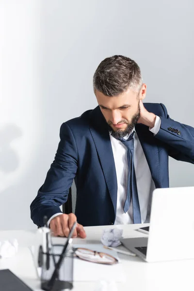 Businessman touching neck near laptop and stationery on blurred foreground in office — Stock Photo