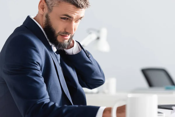 Businessman suffering from neck pain near cup on blurred foreground in office — Stock Photo