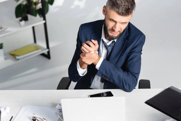 Uomo d'affari che soffre di dolore al braccio mentre è seduto vicino a gadget e carta in primo piano sfocato in ufficio — Foto stock