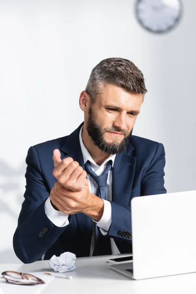 Empresario tocando el brazo mientras sufre de dolor cerca de gadgets en la mesa de trabajo en primer plano borroso en la oficina - foto de stock