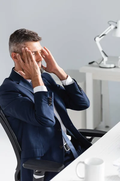 Empresário em desgaste formal tocando a cabeça durante a enxaqueca perto do copo em primeiro plano desfocado no escritório — Fotografia de Stock