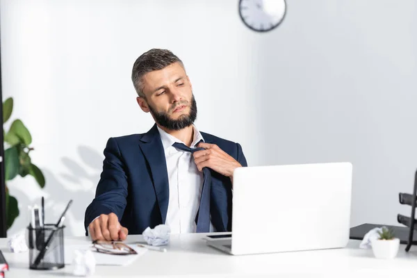 Geschäftsmann berührt Krawatte, während er unter Hitze in der Nähe von Gadgets und Schreibwaren auf verschwommenem Vordergrund leidet — Stockfoto