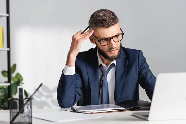 Empresario en anteojos sosteniendo pluma mientras trabaja con papeles y portátil en primer plano borroso en la oficina - foto de stock