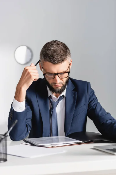 Homem de negócios concentrado segurando caneta enquanto olha para documentos perto do laptop em primeiro plano turvo — Fotografia de Stock