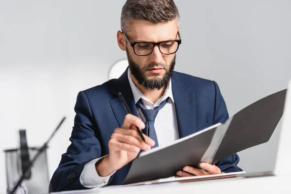 Empresário em óculos segurando pasta de papel e caneta em primeiro plano desfocado no escritório — Fotografia de Stock