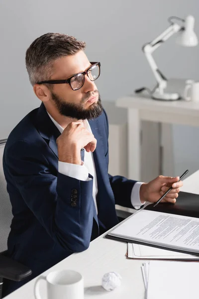 Виснажений бізнесмен тримає ручку біля документів і чашку на розмитому передньому плані в офісі — стокове фото