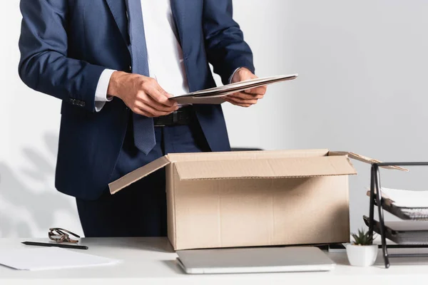 Cropped view of dismissed businessman holding paper folder near laptop and cardboard box on blurred foreground — Stock Photo