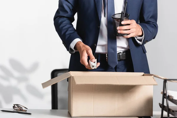 Обрізаний вид звільненого бізнесмена, що тримає канцелярські товари біля картонної коробки на столі в офісі — стокове фото