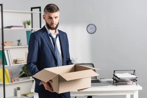 Empresário demitido segurando caixa de papelão com mesa de escritório no fundo embaçado — Fotografia de Stock