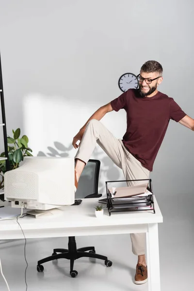 Homem de negócios irritado chutando monitor de computador perto de artigos de papelaria no escritório — Fotografia de Stock