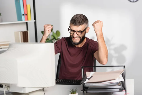 Сердитий бізнесмен з руками в кулаках дивиться на комп'ютер біля паперів на розмитому фоні — стокове фото
