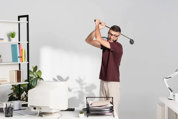 Empresario enojado sosteniendo club de golf cerca de la computadora y documentos en la mesa de oficina - foto de stock