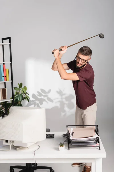 Enojado hombre de negocios con club de golf de pie cerca de la computadora y documentos en primer plano borroso en la oficina - foto de stock