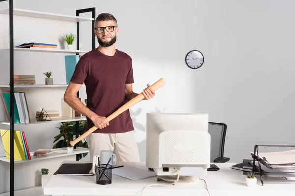 Empresario sosteniendo bate de béisbol mientras está de pie cerca de la computadora, papeles y artículos de papelería en primer plano borroso en la oficina - foto de stock