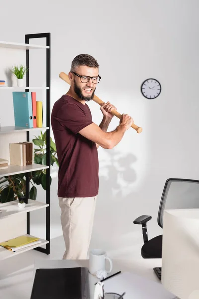 Loco hombre de negocios sosteniendo bate de béisbol cerca de la computadora en primer plano borroso en la oficina - foto de stock