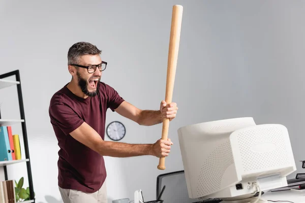 Empresario loco sosteniendo bate de béisbol cerca del monitor de la computadora en primer plano borroso en la oficina - foto de stock