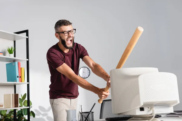 Schreiender Geschäftsmann schlägt mit Baseballschläger auf Computermonitor auf verschwommenem Bürotisch ein — Stockfoto