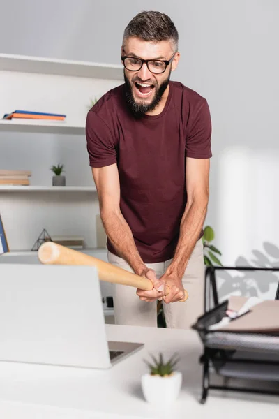 Кричущий бізнесмен побиває ноутбук з бейсбольною битою на розмитому передньому плані в офісі — стокове фото