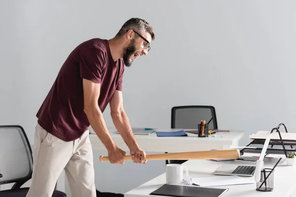 Empresário estressado segurando bastão de beisebol perto de laptop e papelaria na mesa de escritório — Fotografia de Stock