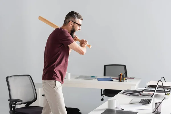 Vue latérale d'un homme d'affaires criant tenant une batte de baseball près d'un ordinateur portable et de la papeterie au bureau — Photo de stock
