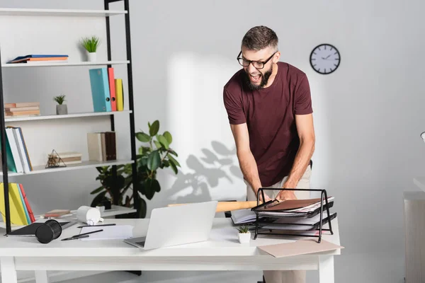 Arrabbiato uomo d'affari che picchia computer portatile con mazza da baseball vicino cancelleria sul tavolo dell'ufficio — Foto stock