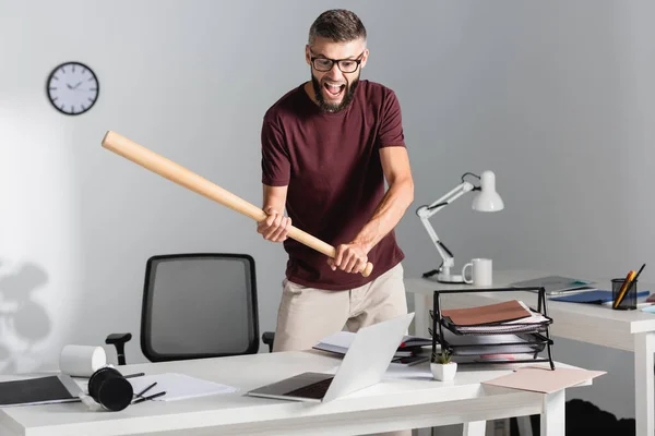 Кричащий бизнесмен, держащий бейсбольную биту возле ноутбука и канцелярских принадлежностей во время нервного срыва в офисе — стоковое фото
