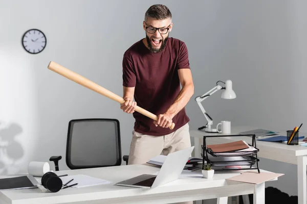 Urlando uomo d'affari in possesso di mazza da baseball vicino computer portatile e cancelleria durante esaurimento nervoso in ufficio — Foto stock