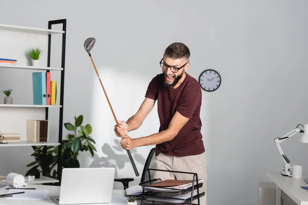 Безумный бизнесмен держит гольф-клуб рядом с ноутбуком и канцелярскими принадлежностями в офисе — стоковое фото