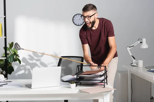 Wütender und schreiender Geschäftsmann hält Golfschläger in der Nähe von Dokumenten und Laptop im Büro — Stockfoto