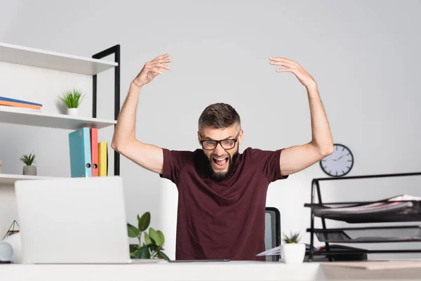 Hombre de negocios enojado gritando cerca de la computadora portátil y artículos de papelería en primer plano borroso en la oficina - foto de stock