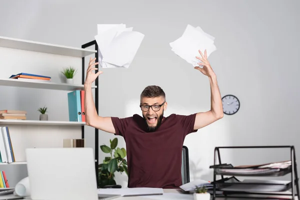 Homem de negócios gritando jogando papéis perto do laptop em primeiro plano desfocado no escritório — Fotografia de Stock