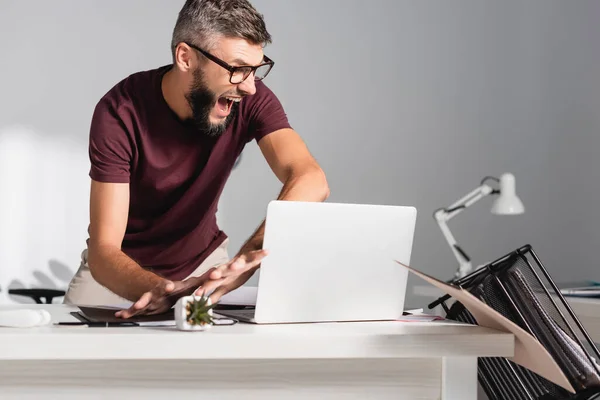 Screaming businessman throwing laptop, papers and stationery from table — Stock Photo