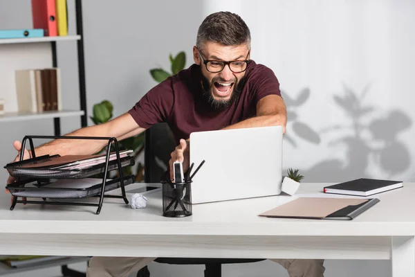 Agresivo hombre de negocios gritando mientras lanza el portátil y los papeles de la mesa en la oficina - foto de stock