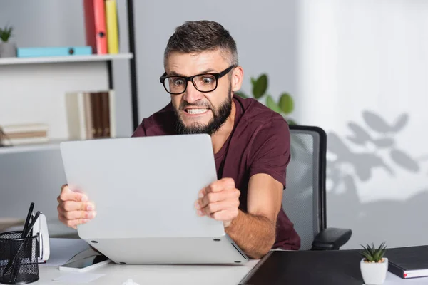 Homme d'affaires fou tenant un ordinateur portable près du dossier papiers sur le premier plan flou dans le bureau — Photo de stock