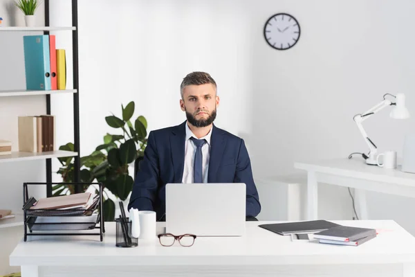 Hombre de negocios mirando la cámara cerca de gadgets y carpetas de papeles en primer plano borroso en la oficina - foto de stock