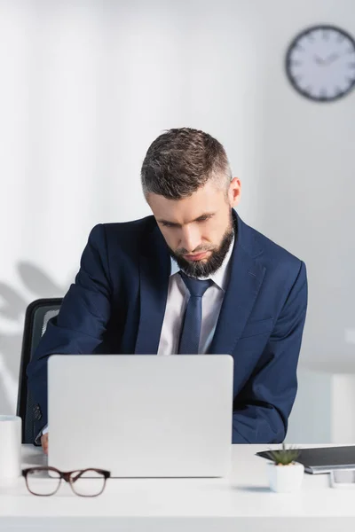 Бизнесмен, использующий ноутбук возле очков на размытом переднем плане в офисе — стоковое фото
